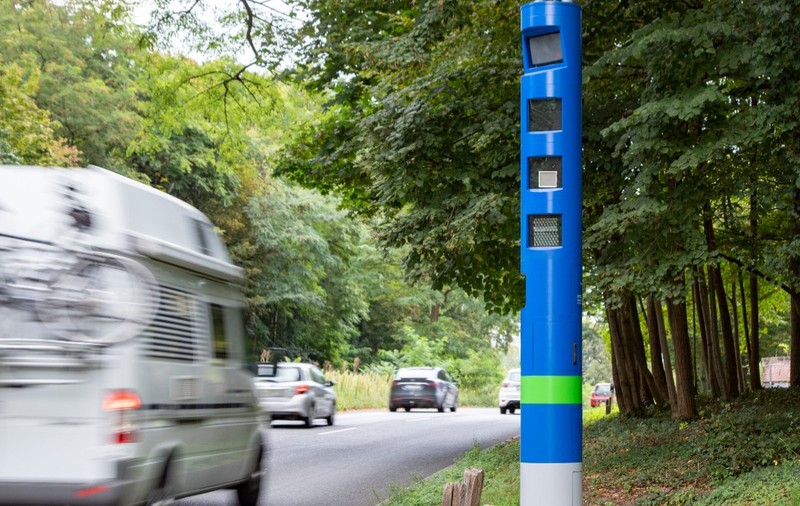 Un stâlp cilindric albastru înalt, prevăzut cu camere de supraveghere și senzori, amplasat pe marginea unei șosele mărginite de copaci în timp ce trec vehicule.