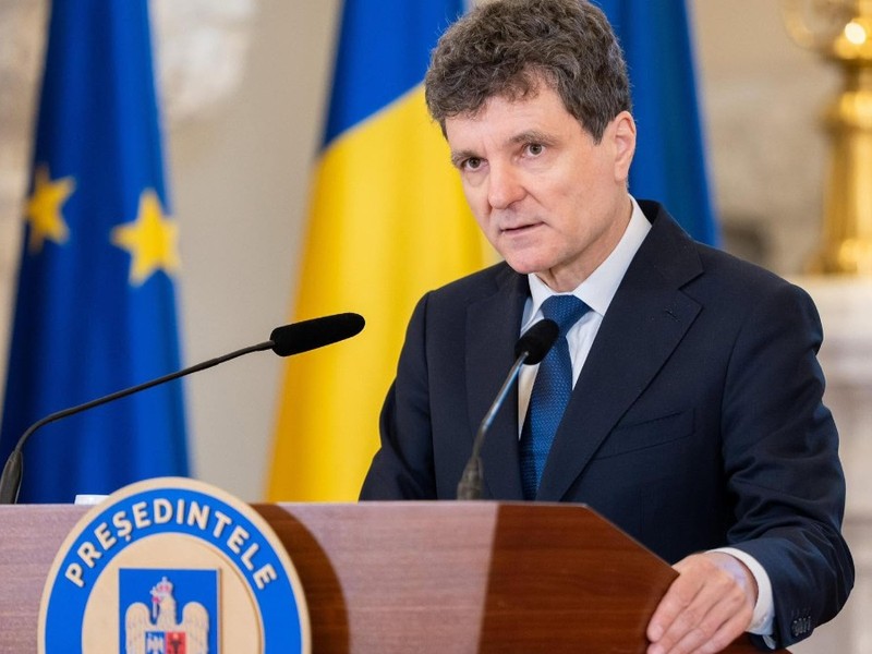 Imaginea îl prezintă pe Nicușor Dan vorbind de la o tribună oficială, având în fundal steagurile României și Uniunii Europene.