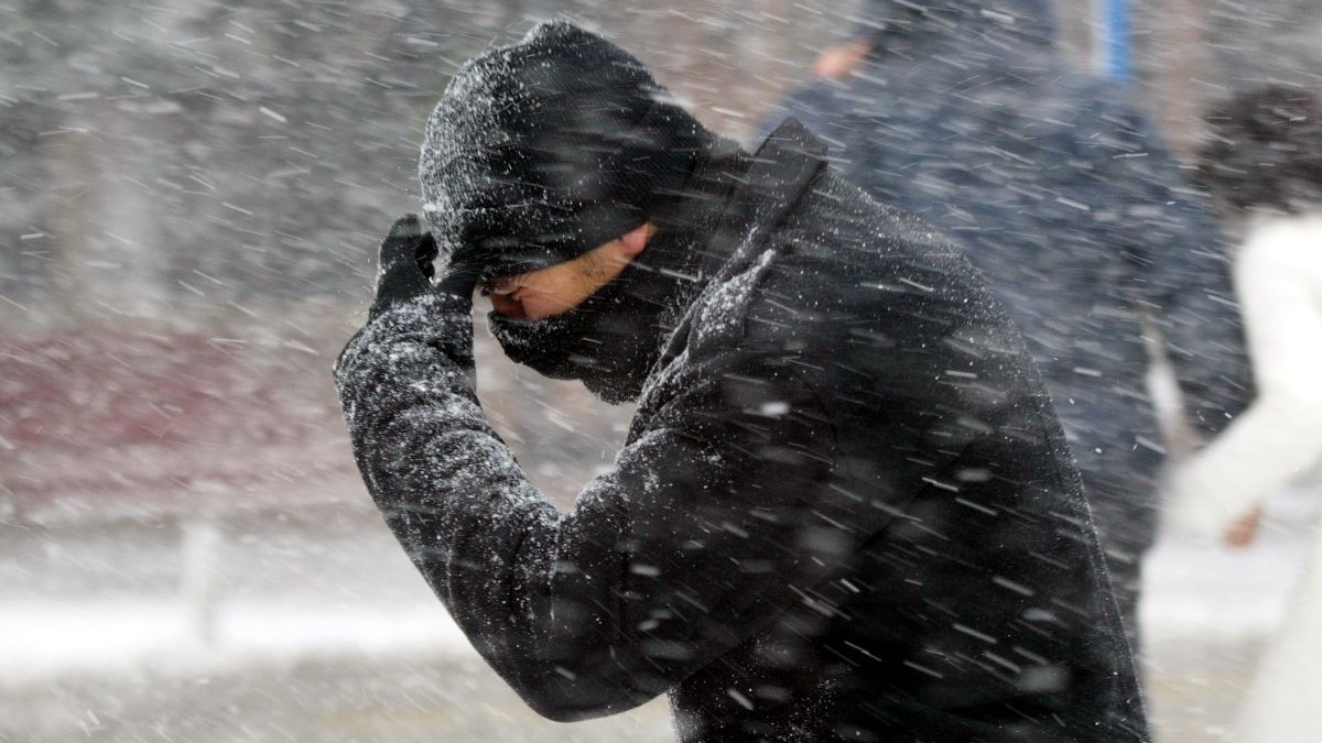 Fotografie cu un bărbat îmbrăcat într-o haină neagră, căciulă și mănuși negre, care își protejează fața de ninsoarea abundentă într-un cadru urban.