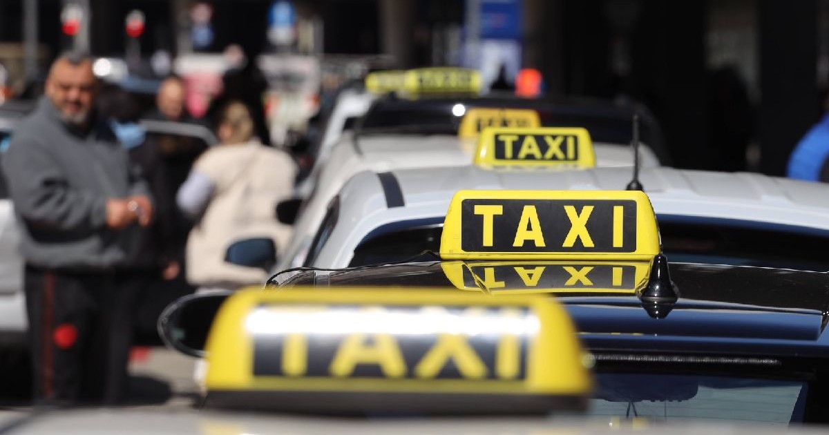 Imaginea arată un șir de taxiuri parcate unul în spatele celuilalt, cu mai multe persoane estompate în fundal.
