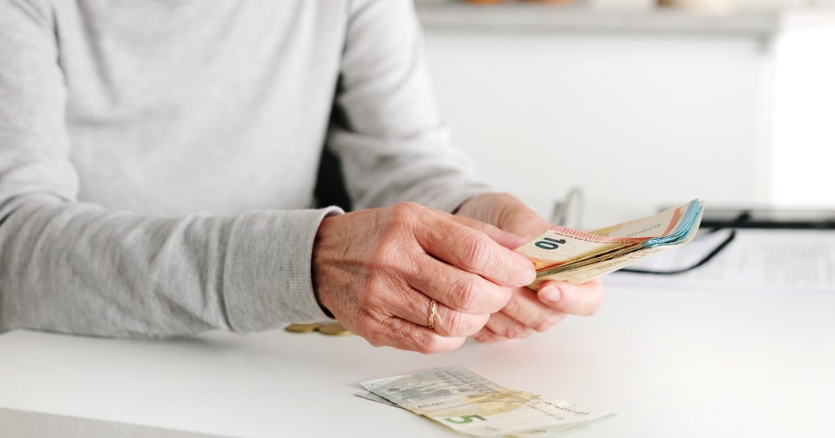 Imaginea arată o persoană în vârstă care numără bancnote euro la o masă, într-un cadru de acasă. Fotografia sugerează preocupările legate de pensie, venituri și gestionarea cheltuielilor de zi cu zi.