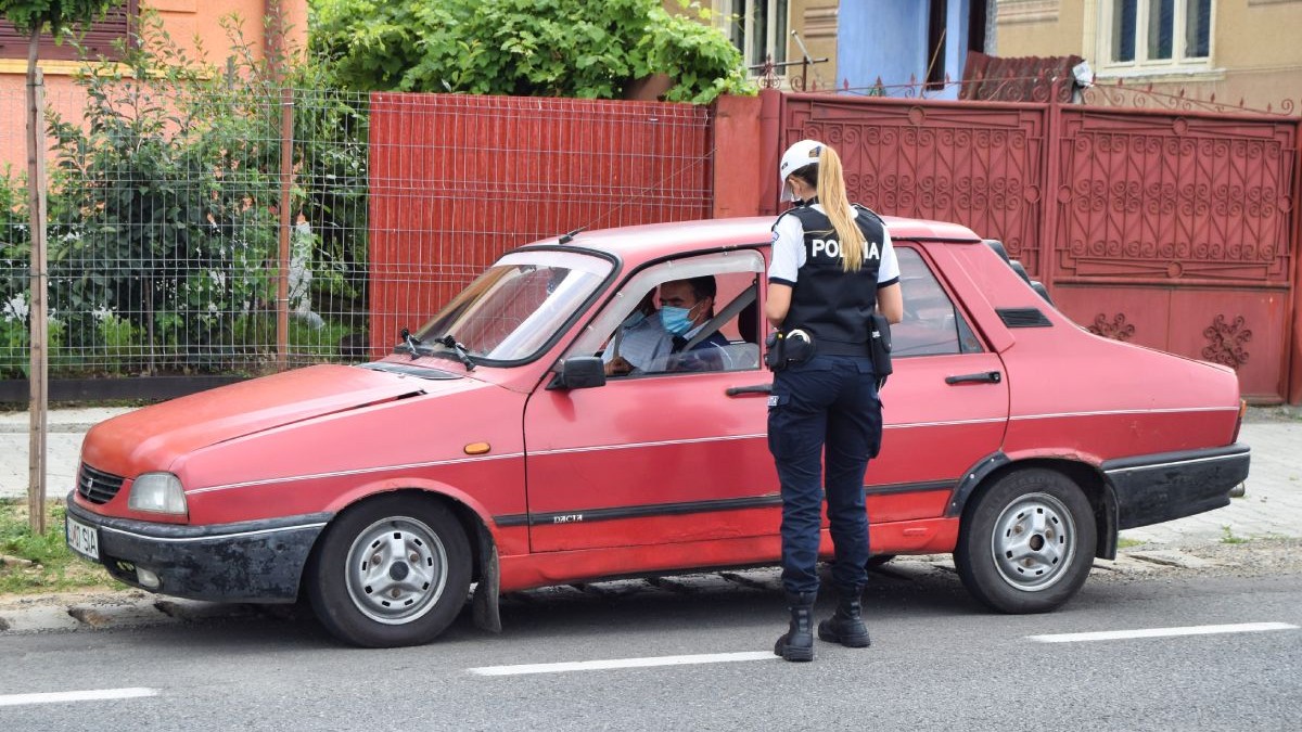 O polițistă îmbrăcată în uniformă controlează actele unui șofer care conduce o Dacia 1300 de culoare roșie