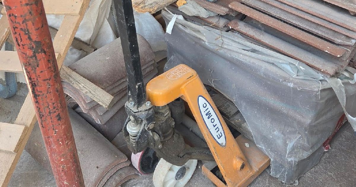 Fotografie cu un șantier aglomerat, cu o țeavă metalică neagră, o clemă galbenă „Clawhamer” și diverse materiale de construcție împrăștiate.