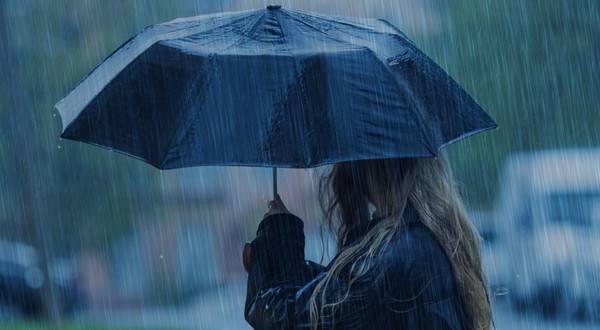 O persoană cu părul lung și blond, privită din lateral, care ține o umbrelă neagră deschisă pe o ploaie torențială.