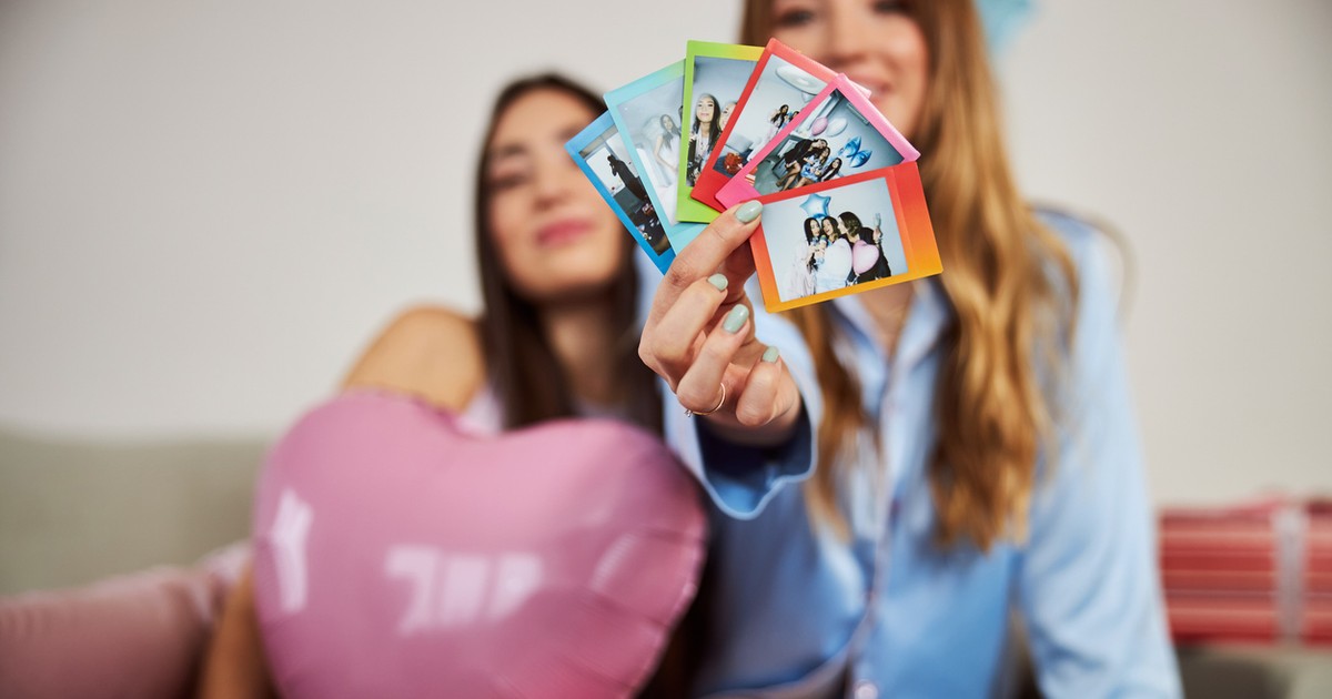 Doua fete, una bruneta si una blonda. Cea bruneta este in stanga si tine in brate un balon roz in forma de inima, cea blonda sta in drepata si tine in mana poze instant