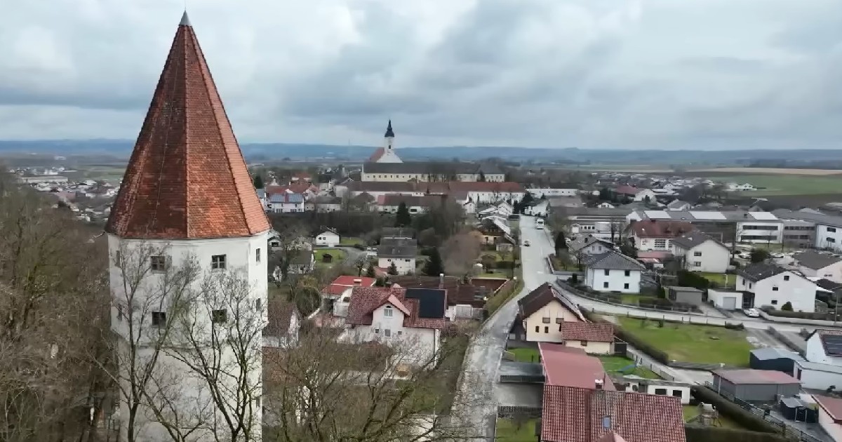 Imaginea oferă o vedere panoramică asupra unui orășel pitoresc, evidențiind un turn alb cu acoperiș conic din țiglă roșie în prim-plan.