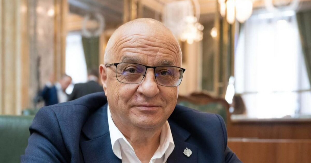 Imaginea îl surprinde pe un bărbat îmbrăcat elegant, purtând un sacou bleumarin și o cămașă albă. Poartă ochelari cu rame subțiri și are o expresie serioasă, dar calmă. Fundalul pare a fi o încăpere oficială sau o sală de ședințe, decorată cu candelabre și elemente elegante, sugerând un cadru instituțional sau politic.