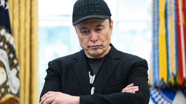 Elon Musk stând cu brațele încrucișate și privind în jos, îmbrăcat într-o jachetă neagră și purtând o șapcă neagră inscripționată cu textul „DOGE”.