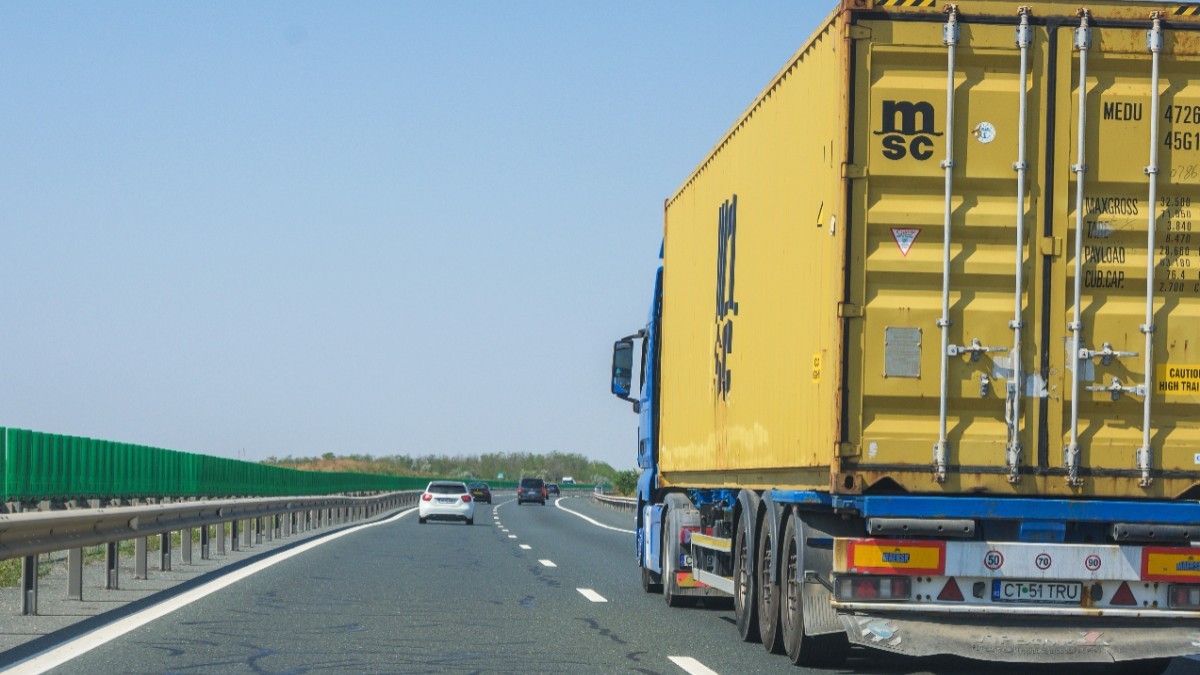 Imaginea surprinde un camion albastru ce transportă un container galben, rulând pe o autostradă alături de alte autoturisme, sub un cer senin.