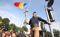 Un bărbat cu barbă și brâu din piele, purtând un toiag decorat și un steag al României, urcat la înălțime deasupra unei mulțimi de protestatari.