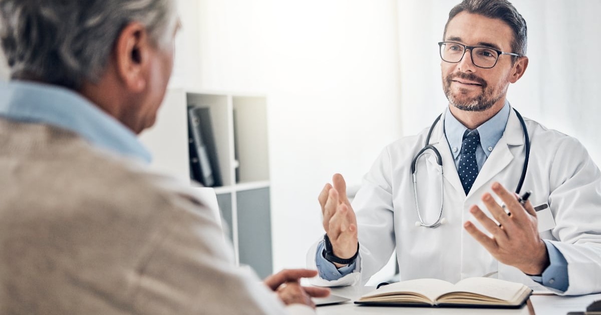 Imaginea surprinde un medic în timpul unei consultații, discutând cu un pacient aflat în fața sa. Doctorul pare atent și explică detalii, într-un cadru medical modern și luminos.