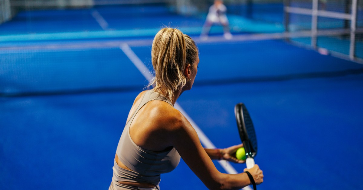 O femeie se pregătește să lovească mingea în timpul unui meci de padel, concentrată pe joc. Terenul albastru și poziția ei sugerează un moment dinamic, surprins în timpul unei acțiuni sportive.
