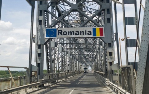 Podul Giurgiu-Ruse, podul Prieteniei, văzut dinspre Giurgiu. Pe șosea se află un TIR alb, iar deasupra ramei metalice ale podului se observă un semn de circulație cu textul „România”, steagul Uniunii Europene și steagul României