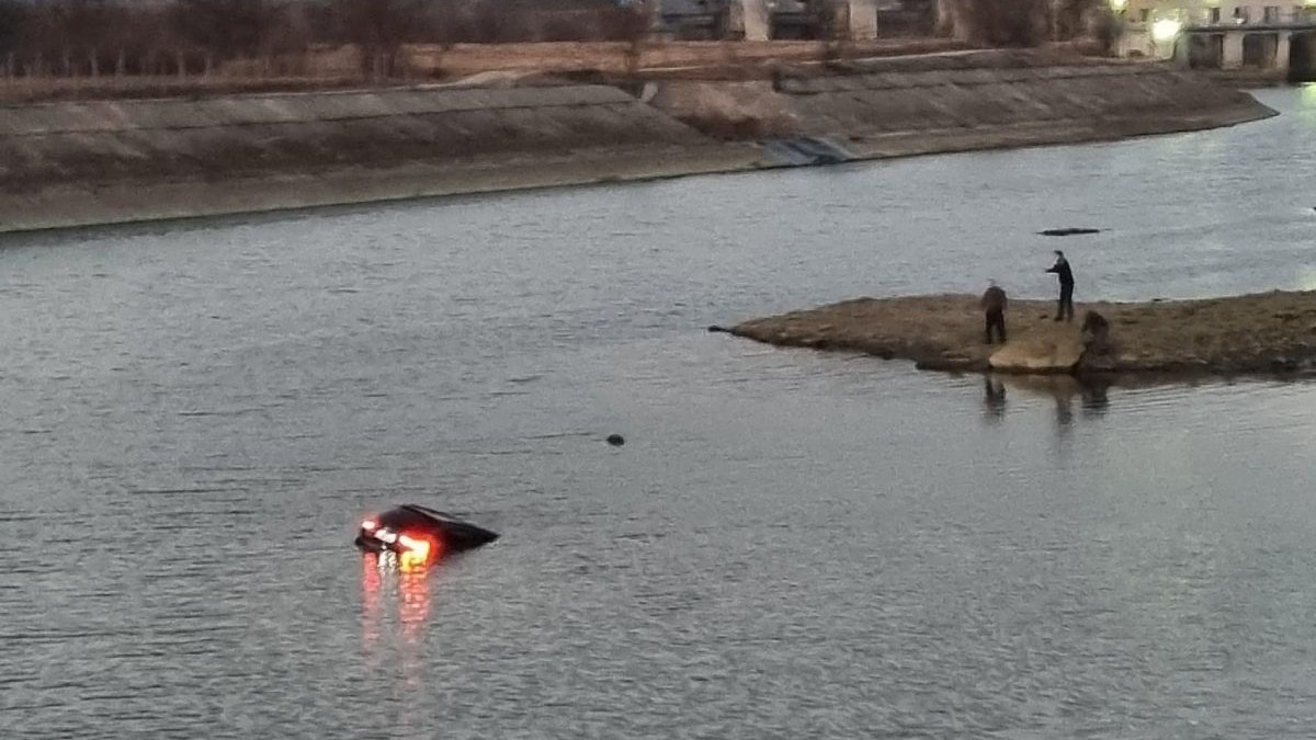 Doi oameni pe malul râului Olt, în timp ce privesc către mașina scufundată în apă căreia încă i se mai văd stopurile aprinse