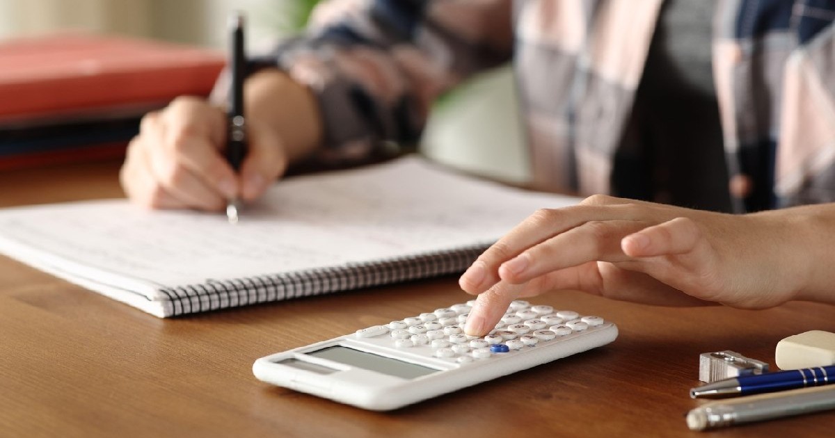 Mâinile unui student care scrie. Acesta folosește un calculator, pe o masă de lemn.