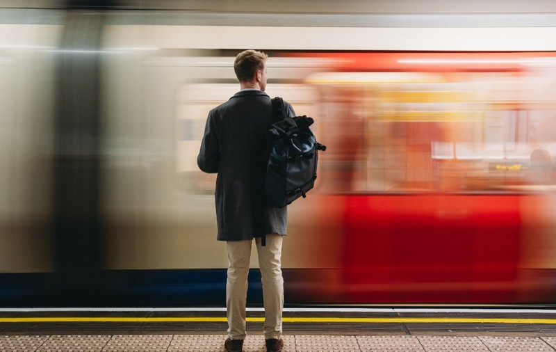 Un bărbat cu rucsac în spate, văzut din spate pe peronul unei stații de metrou, în timp ce prin fața lui trece un tren în viteză.