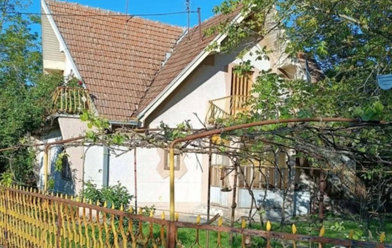O casă cu etaj și mansardă, într-o curte plină de trandafiri. Are gardul galben și acoperișul din țiglă roșie