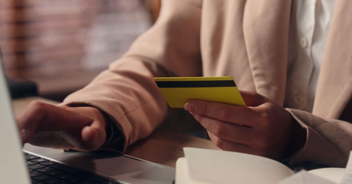 cadru strans mainile unei femei care sta la laptop, mana dreapta pe tastatura, in mana stanga tine un card de credit