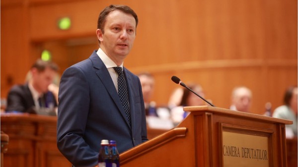 Europarlamentarul Siegfried Mureșan vorbind de la tribuna Parlamentului, îmbrăcat într-un costum albastru și cravată cu picățele.   