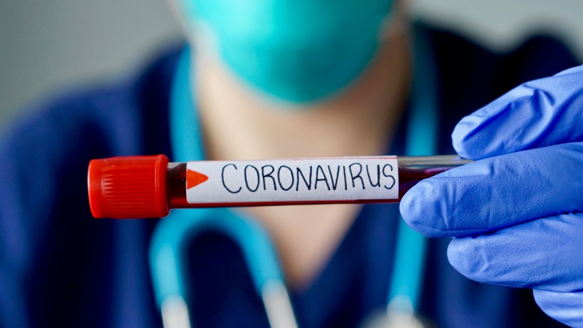 ”Noul normal”. Cum va arăta economia după pandemie. Doctor îmbrăcat în combinezon, cu stetoscop în jurul gâtului și eprubetă în mână, pe care scrie Coronavirus
