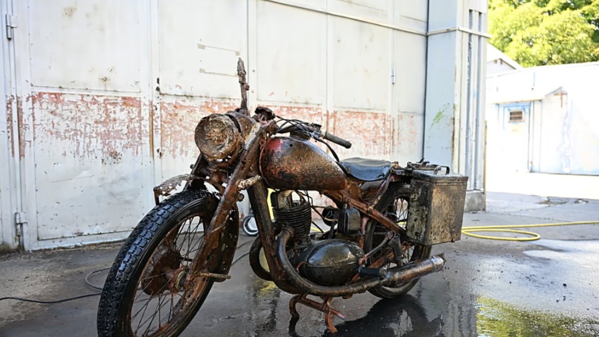 În imagine apare o motocicletă veche, puternic ruginită, care prezintă urme vizibile de uzură după o perioadă îndelungată de degradare. Vehiculul este surprins în aer liber, în fața unei clădiri industriale, după ce pare să fi fost curățat sau spălat.
