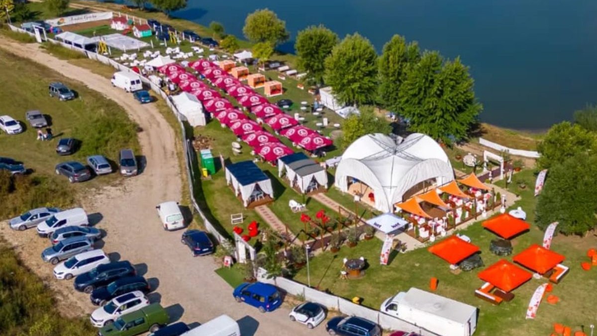 Imagine aeriană cu localul Arruba Garden din localitatea Paleu, Bihor, pe marginea unui lac, cu corturi și umbrele, și mai multe mașini parcate într-o parcare și pe un drum de acces