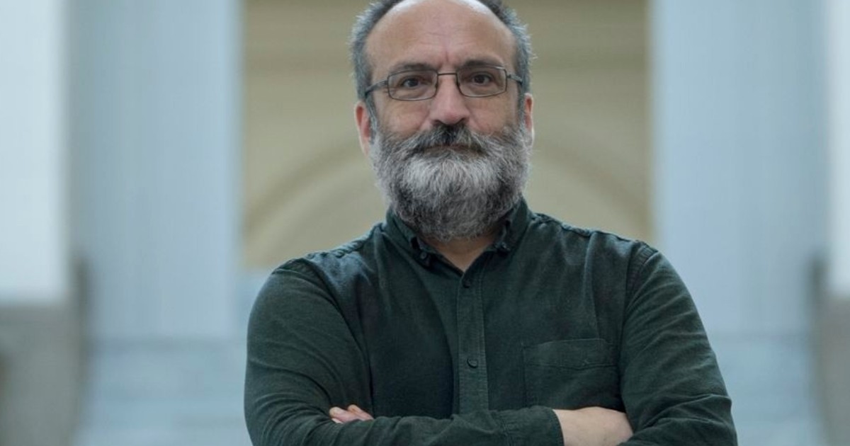 În imagine apare un bărbat cu barbă și ochelari, îmbrăcat într-o cămașă închisă la culoare, fotografiat într-un spațiu interior sobru.