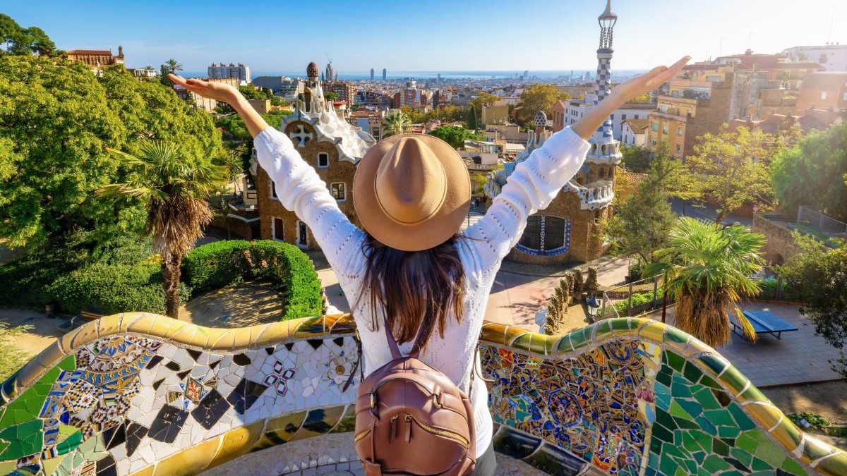 O femeie , turista, cu spatele, cu o palarie pe cap si ghiozdan in spate, cu mainile ridicate, larg deschise, in Park Güell, Barcelona, Spaina, intr-o zi insorita