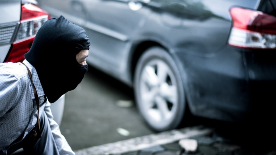 Un bărbat purtând o cagulă neagră, ghemuit discret lângă un autoturism parcat.   