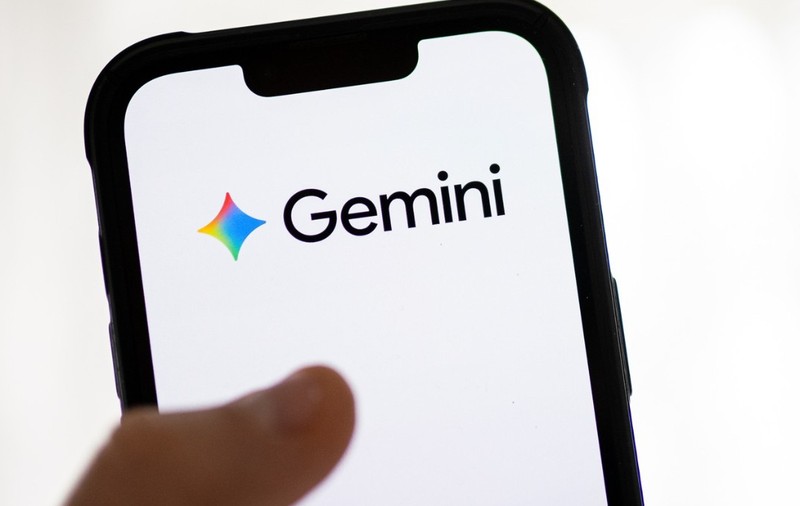 Imaginea prezintă un smartphone ținut în mână, pe al cărui ecran este afișat clar logo-ul și numele "Gemini". Sigla este reprezentată de o formă colorată (roșu, galben, albastru) alături de numele scris cu litere negre pe fundal alb, sugerând o aplicație sau un serviciu digital.