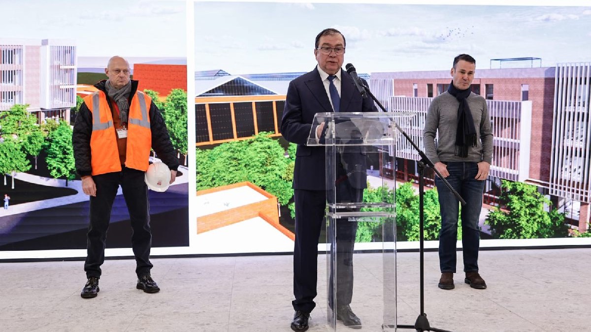 Un constructor cu casca de protectie in mana si vsta reflectorizanta in picioare in stanga, pe miljloc un barbat in costum negru vorbind la microfon, rectorul UMFCD, Viorel Jinga si in dreapta putin mai in spate primarul sectorului 3 Bucuresti Robert Negoita cu mainile in buzunare in spatele lor backgounf cu centrul de excelenta pentru medici- training