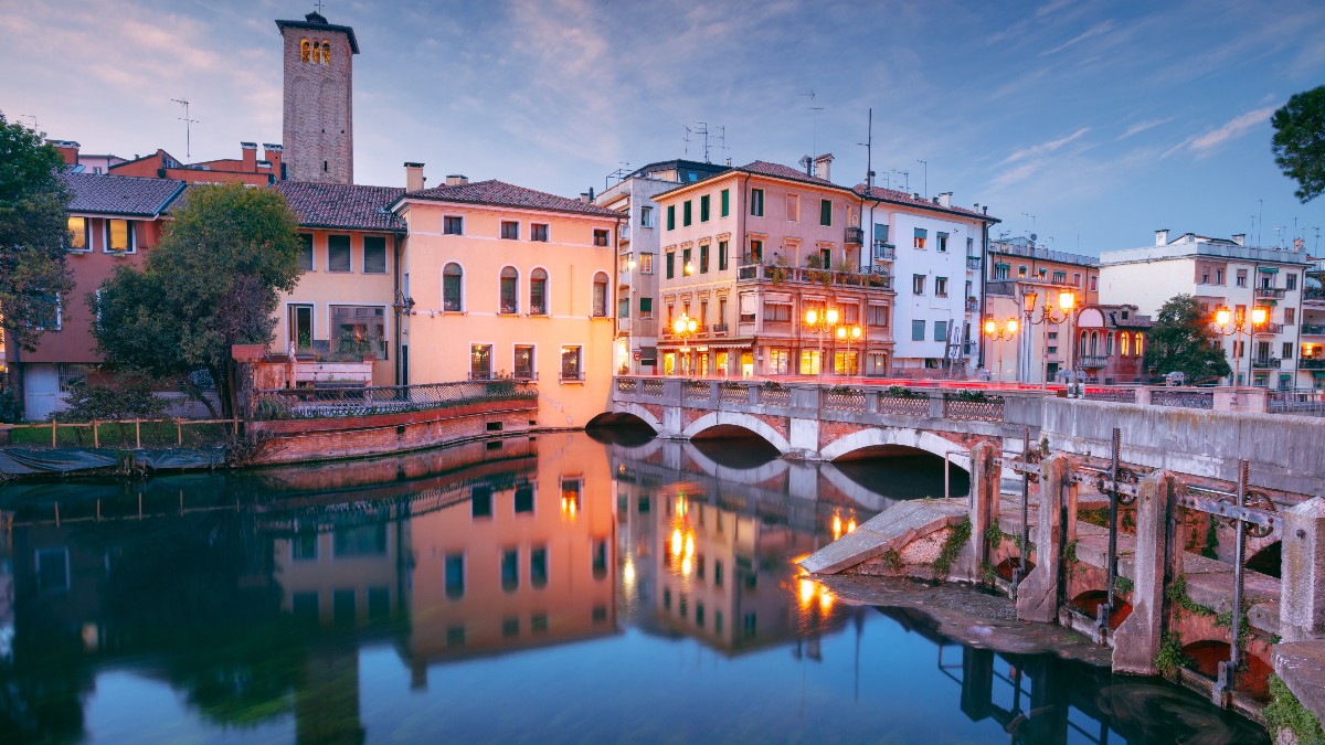 Imaginea surprinde un peisaj urban de seară din Treviso, Italia, evidențiind clădiri vechi cu fațade colorate și acoperișuri din țiglă, iluminate de lumini calde. Clădirile se reflectă în apa unui canal traversat de un pod arcuit, iar în fundal se înalță un turn înalt.