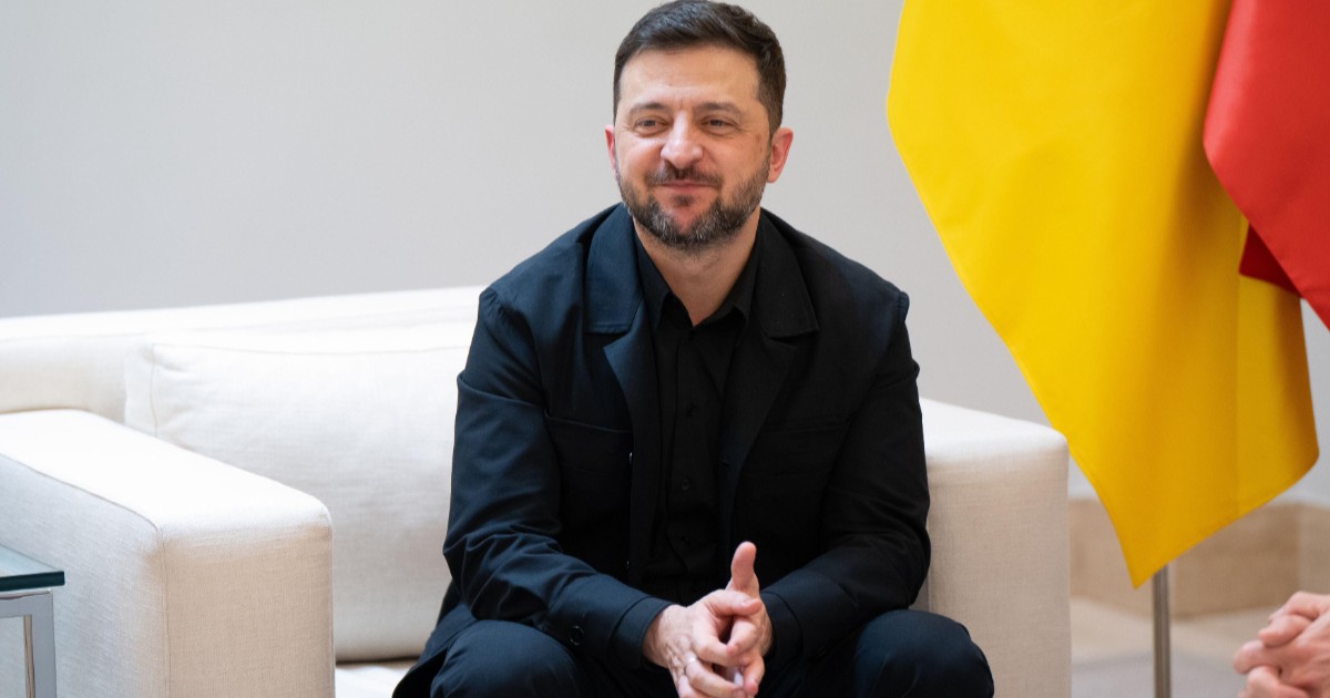 Președintele Volodimir Zelenski, îmbrăcat în negru, pe o canapea albă, în timp ce își ține mâinile încrucișate și zâmbește