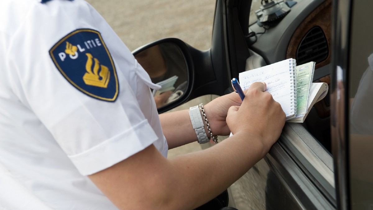 Imaginea, un prim-plan realizat printr-o fereastră de mașină, arată un ofițer de poliție olandez (pe baza emblemei "POLITIE") în uniformă albă. Ofițerul scrie cu un pix într-un carnet spiralat, sugerând momentul întocmirii unui proces-verbal sau luarea de notițe.