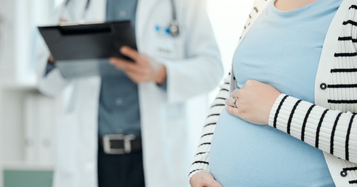 O femeie însărcinată își ține mâna pe burtă, în timp ce un medic se află în fundal, analizând documente. Imaginea sugerează un control medical și o situație care necesită atenție și îngrijire de specialitate.