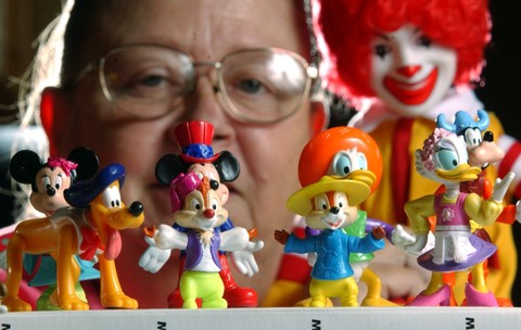 Cinci jucării de la McDonald's, cu figurine Disney, așezate pe o măsuță. În spatele lor se vede o femeie cu ochelari