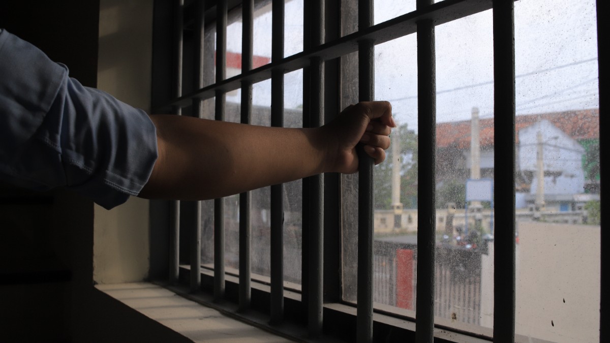 Un bărbat care stă în închisoare și se uită pe fereastră, afară este înnorat, iar el se ține cu mâinile de gratiile închisorii