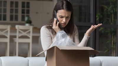 Cum depui o reclamaţie la Protecţia consumatorului – ANPC – O femeie, care stă pe canapea și ține o cutie în brațe, vorbește la telefon și pare nemulțumită de produsul primit