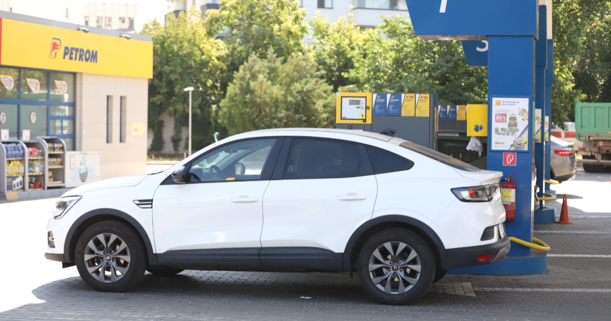 Imaginea prezintă o mașină albă oprită la o pompă dintr-o stație de carburanți Petrom.