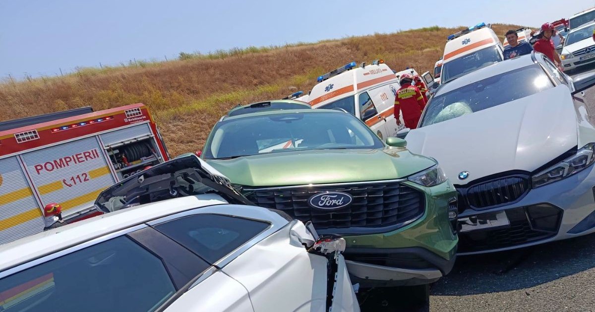 accident rutier în lanț pe o șosea mărginită de un taluz de pământ acoperit cu vegetație. În prim-plan este înfățișată o serie de autoturisme puternic avariate și încastrate unele în altele, având elemente ale caroseriei contorsionate, faruri sparte și bare de protecție smulse în urma coliziunilor. În spatele acestui carambol sunt staționate mai multe vehicule de urgență, printre care o autospecială de pompieri cu compartimente de echipament deschise și mai multe ambulanțe. În spațiul dintre mașinile avariate și autospeciale se află pompieri și paramedici în uniforme de intervenție, alături de martori, evaluând pagubele și desfășurând operațiuni de salvare.