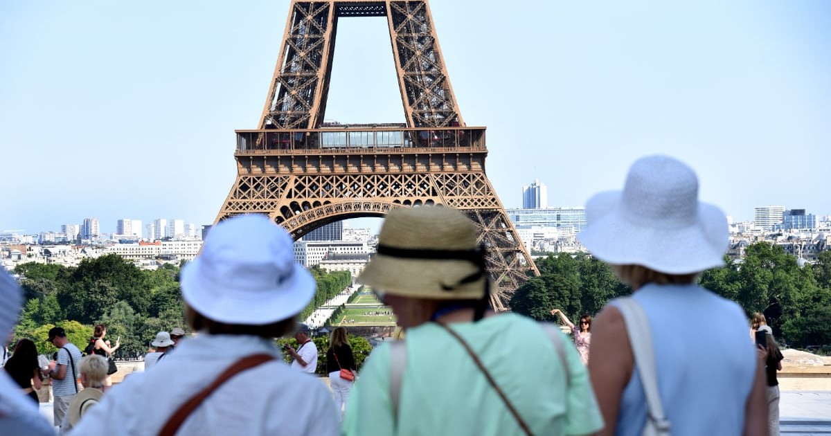 În prim-plan se află mai mulți turiști care admiră și fotografiază Turnul Eiffel, unul dintre cele mai cunoscute simboluri ale Parisului. Imaginea surprinde atmosfera unei zile însorite în capitala Franței, cu numeroși vizitatori adunați în zona de belvedere.
