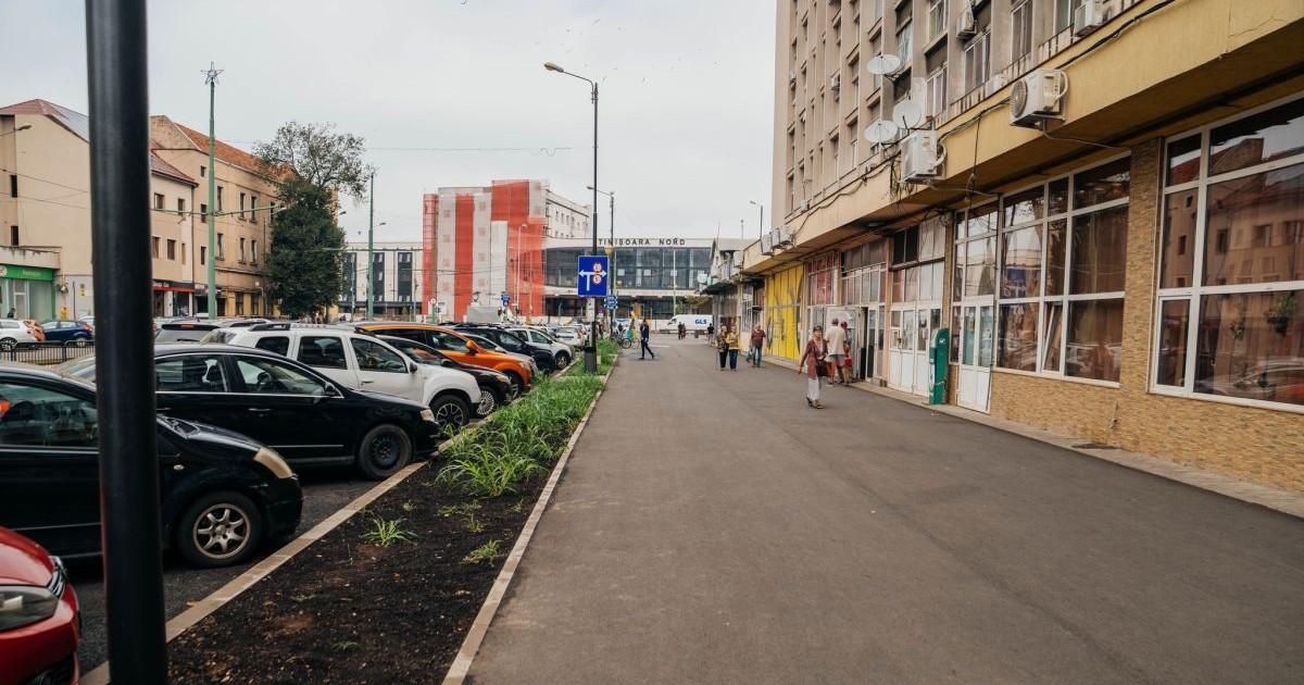 Mai multe pesoane circulă pe un trotur situat lângă un bloc care are magazine și o parcare la parter, în Timișoara, septembrie 2025