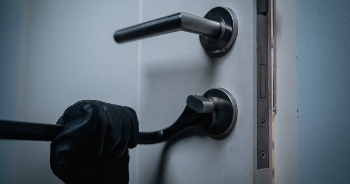 Un bărbat care poartă o mănușă neagră folosește o rangă pentru a forța încuietoarea unei uși albe de apartament.