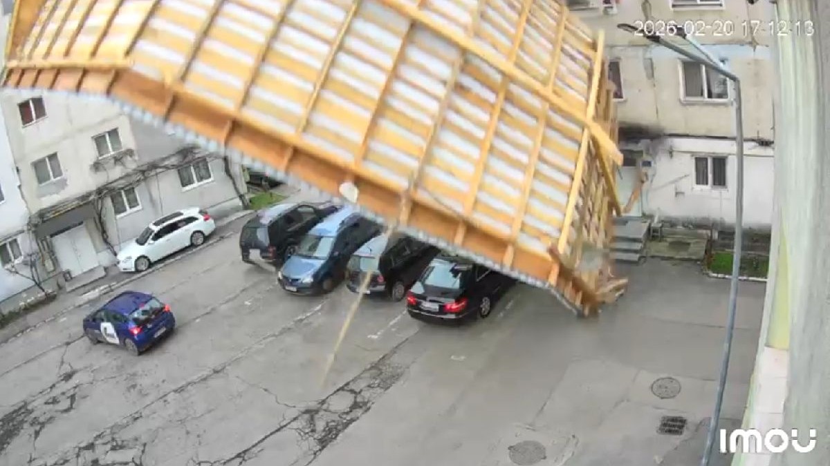O cameră de supraveghere surprinde momentul prăbușirii unui acoperiș de pe un bloc înconjurat de mașini parcate și clădiri adiacente.