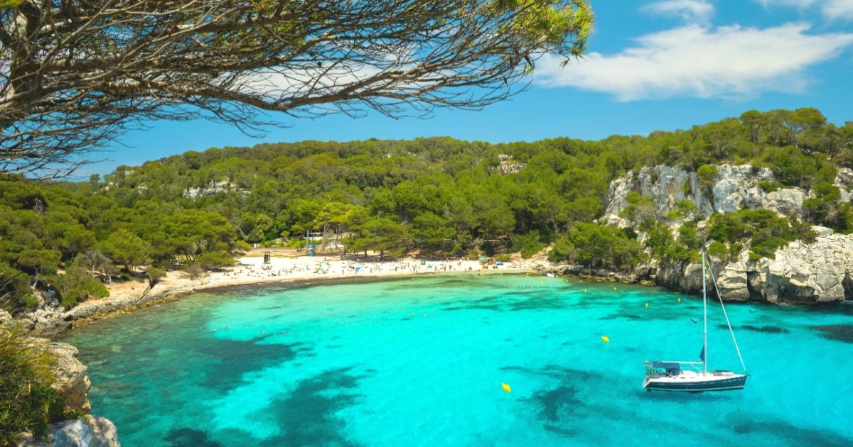 Vedere panoramică a plajei Cala Macarella din Menorca, cu apă turcoaz și o barcă cu pânze ancorată.