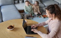 mamă tânără, așezată la o masă, folosind laptopul în timp ce lucrează de acasă cu doi copii jucându-se în fundal, langa o canapea gri