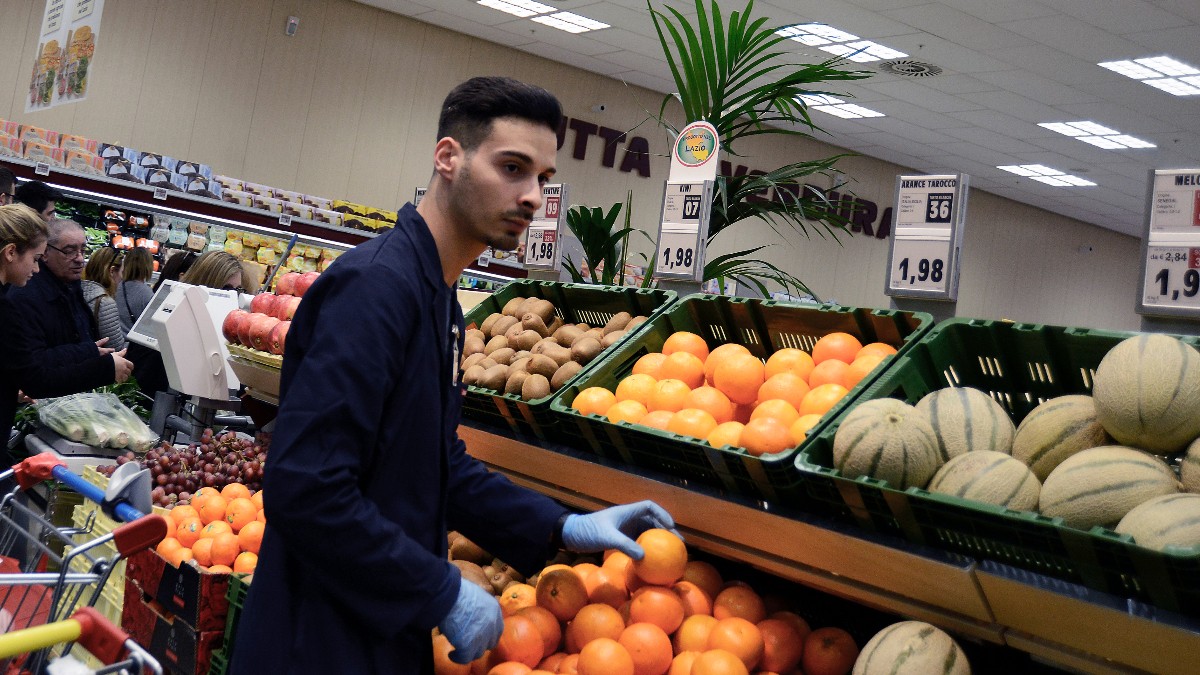 Un tânăr angajat purtând mănuși de protecție aranjează portocale la raionul de fructe și legume dintr-un supermarket.