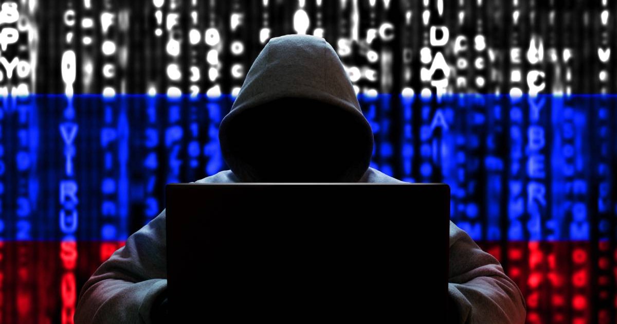 Un spion hacker îmbrăcat în hanorac și cu laptopul în brațe în timp ce pe fundal se vede steagul Rusiei.
