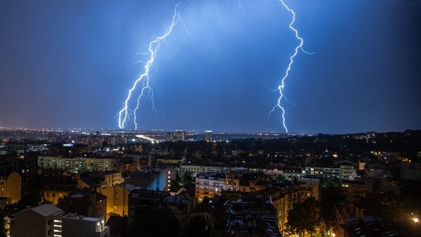 Furtună cu fulgere și cer înnorat peste un oraș, cu două fulgere care lovesc pământul în fundal