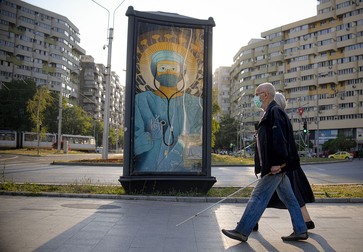 Un cuplu trece pe lângă un panou publicitar cu o ilustrație care înfățișează un medic în echipament de protecție, realizată într-un stil inspirat din icoane religioase ortodoxe, în București, miercuri, 29 aprilie 2020. Foto Hepta